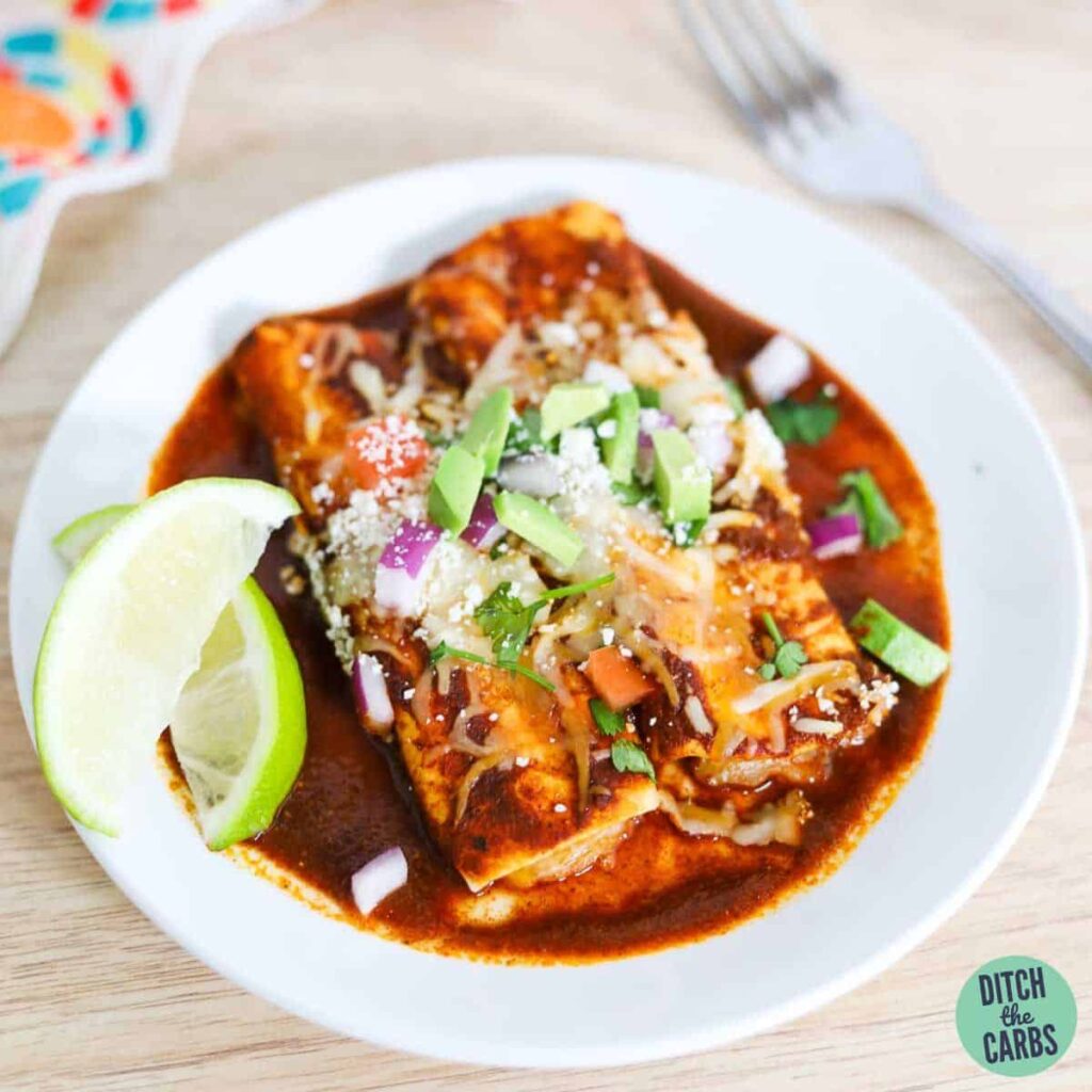 Two keto enchiladas on a white plate topped with onions, tomatoes, cilantro, and avocado.