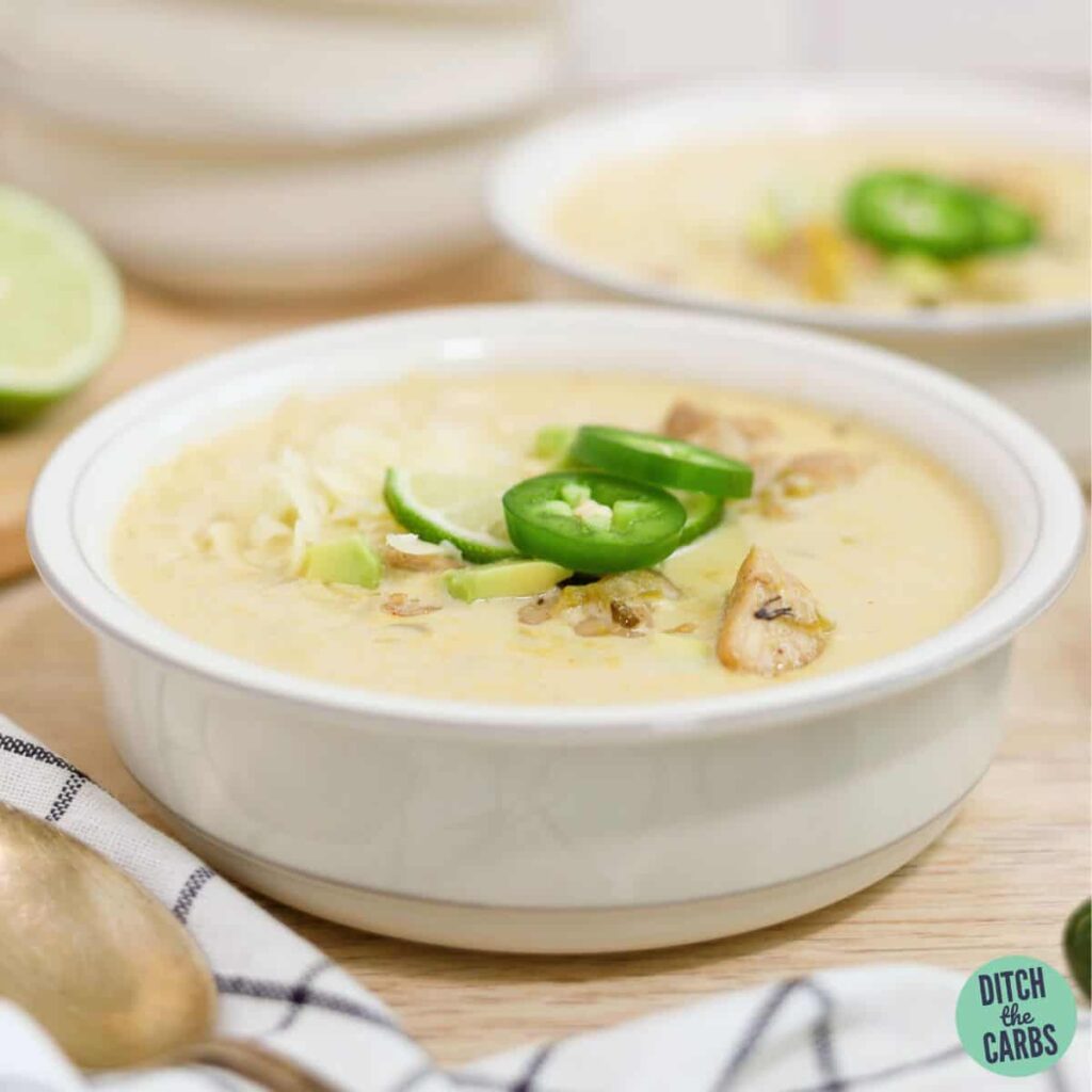 Two bowls of keto white chicken chili on the counter topped with shredded cheese, sliced jalapenos, and a lime wedge.